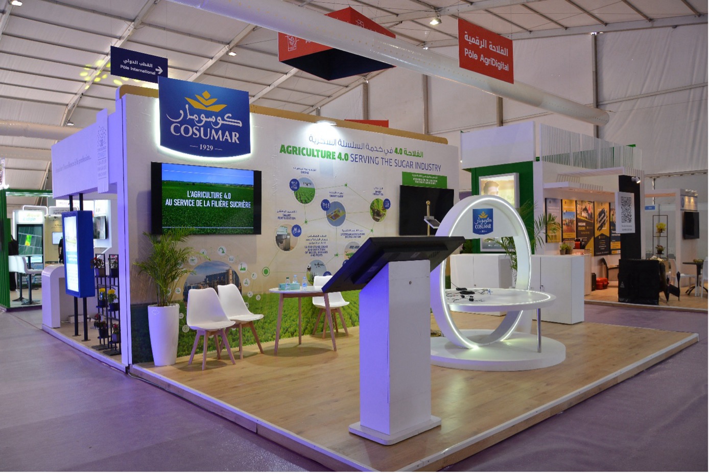 img-cosumar-siam1 | MCG24 | موقع إلكتروني مغربي شامل متجدد على مدار الساعة COSUMAR exhibition booth featuring a large blue logo sign, a circular white display ring, and a wall exhibit about Agriculture 4.0 on a wooden platform.”,