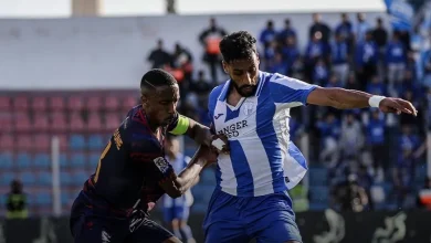 Two soccer players clash for ball; one in blue-and-white stripes shields the ball as the other in dark kit pressures him on a sunny field.