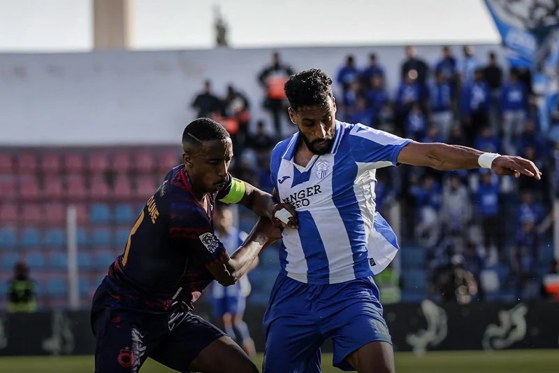 Two soccer players clash for ball; one in blue-and-white stripes shields the ball as the other in dark kit pressures him on a sunny field.