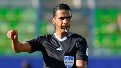 FIFA referee in a black kit on a soccer field, blowing a whistle and signaling with his arm raised.