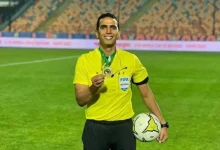 Soccer referee in a bright yellow uniform on the field, holding a medal and a ball with a FIFA badge visible in his chest pocket.