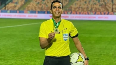Soccer referee in a bright yellow uniform on the field, holding a medal and a ball with a FIFA badge visible in his chest pocket.