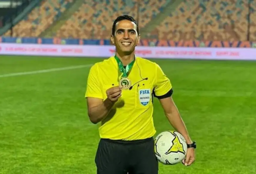 Soccer referee in a bright yellow uniform on the field, holding a medal and a ball with a FIFA badge visible in his chest pocket.