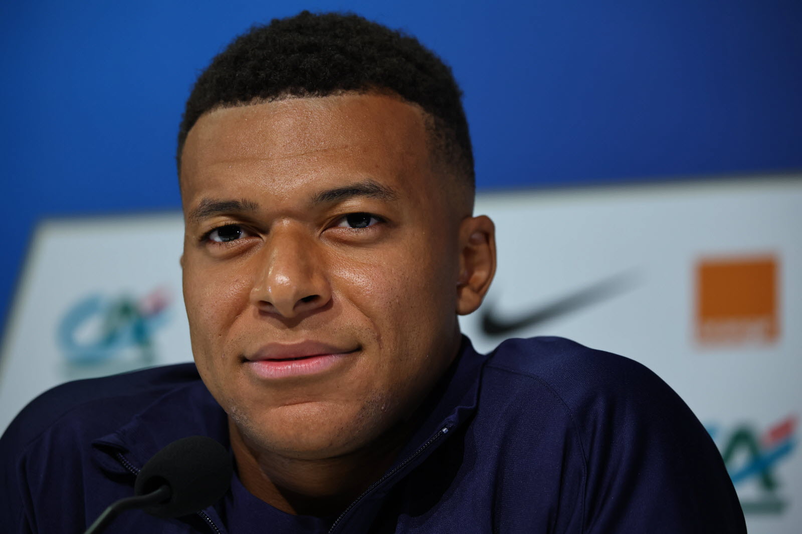 Close-up of a young man speaking at a press conference, microphone in foreground and blue backdrop behind him.