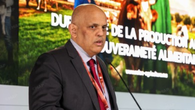 Bald man in a dark suit and red tie speaking at a podium, with a large green banner and text behind him at an event.