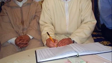 Man in a light beige traditional outfit signs a document with a pencil as others sit nearby at a table.