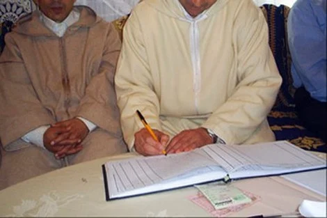 Man in a light beige traditional outfit signs a document with a pencil as others sit nearby at a table.