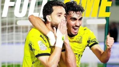 Two male soccer players in bright yellow uniforms celebrate a goal, hugging and smiling near the goal net on the field.