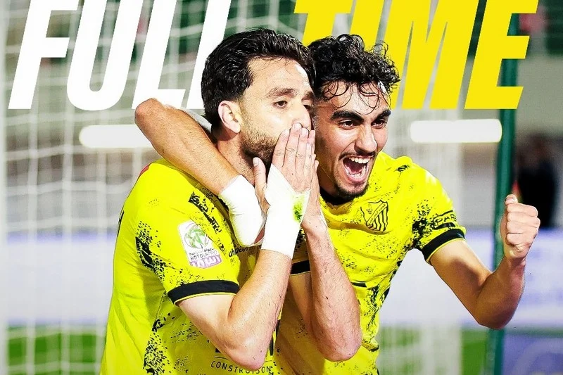Two male soccer players in bright yellow uniforms celebrate a goal, hugging and smiling near the goal net on the field.