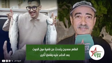Young man with glasses holds two large fish beside a man in a cap giving a TV interview; Arabic caption overlay on screen.