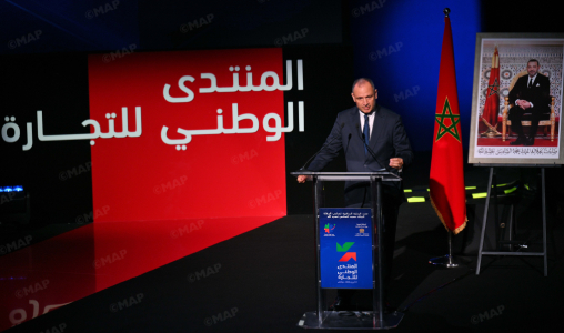 Man at a podium delivering a speech on a stage with a red backdrop displaying Arabic text, next to the Moroccan flag and a framed portrait on an easel.