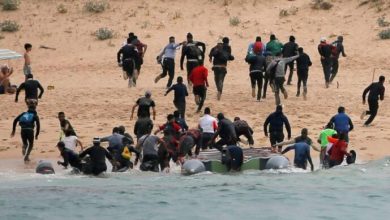 Group of people running from a small inflatable boat onto a sandy beach, some wading in shallow water near the shore.