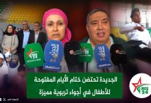 Press conference at a stadium with microphones in the foreground; a woman in a pink headscarf and a man speak while officials and children stand behind them.