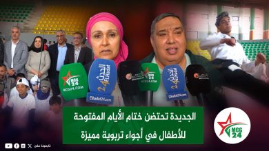 Press conference at a stadium with microphones in the foreground; a woman in a pink headscarf and a man speak while officials and children stand behind them.