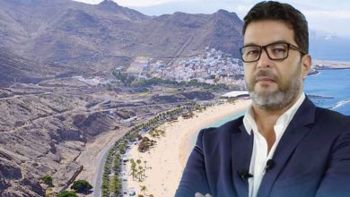 Portrait of a man in a suit and glasses with a coastal town and sandy beach behind him.