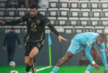 Two soccer players chase the ball on a green field during a match, one in a black kit and the other in light blue.