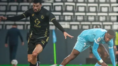 Two soccer players chase the ball on a green field during a match, one in a black kit and the other in light blue.