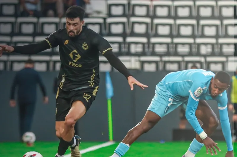 Two soccer players chase the ball on a green field during a match, one in a black kit and the other in light blue.