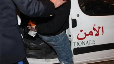 Two men stand near a white van with red Arabic text, one helping the other into the vehicle at night.