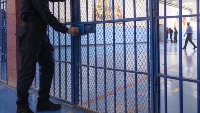 Security guard in black uniform opening a blue metal gate to an indoor facility, with people walking in the background.