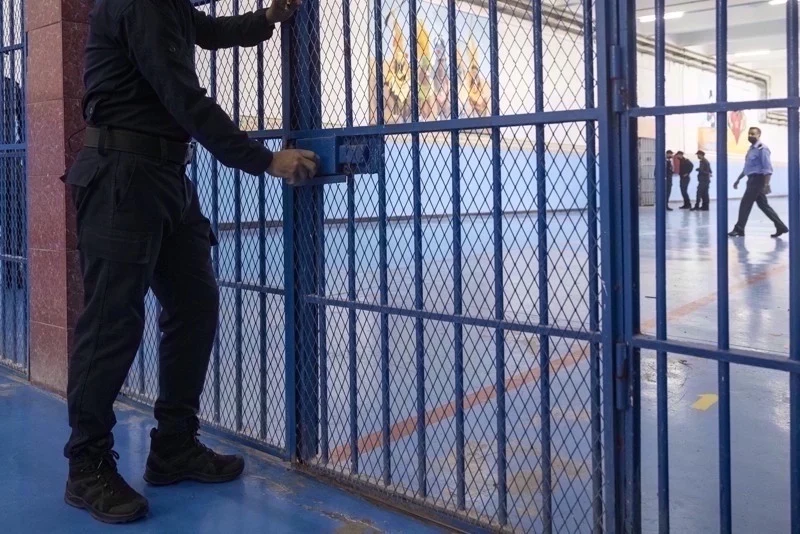 Security guard in black uniform opening a blue metal gate to an indoor facility, with people walking in the background.