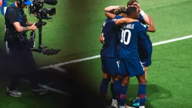 Soccer players in blue celebrate with a group hug on the green field as a cameraman films nearby.