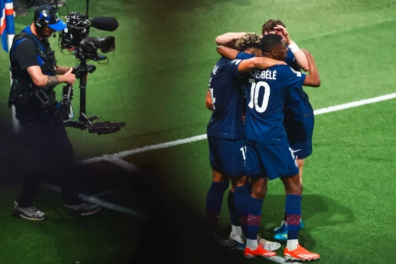 Soccer players in blue celebrate with a group hug on the green field as a cameraman films nearby.