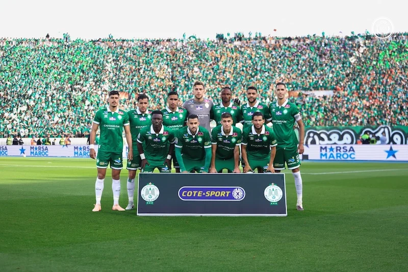 Soccer team in green jerseys posing for a group photo on the field with a banner reading COTE-SPORT and a packed green-clad stadium behind them **(informative image).