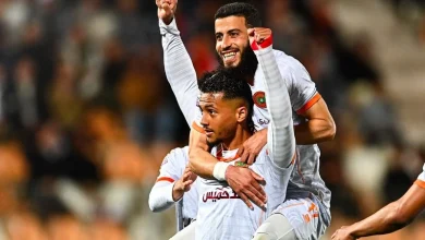Two soccer players in white jerseys celebrate a goal, one lifting the other in jubilation on the field.