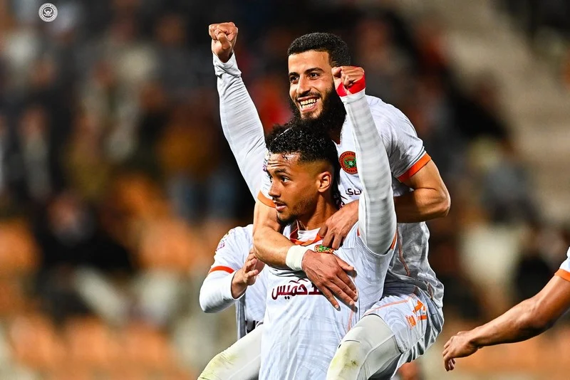Two soccer players in white jerseys celebrate a goal, one lifting the other in jubilation on the field.