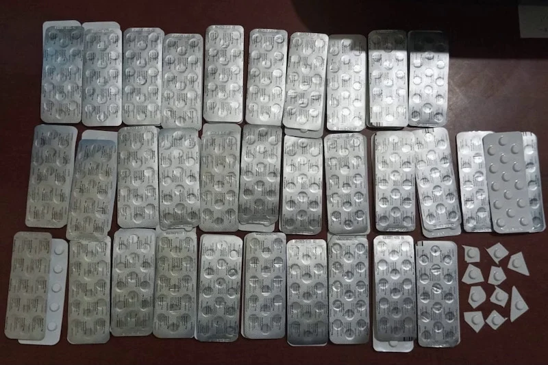 Numerous silver blister packs with round white pills laid out in rows on a dark wooden table.