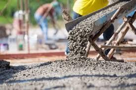 Worker pours wet concrete from a wheelbarrow at a construction site, with other workers and scaffolding in the background.