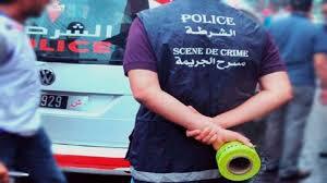 Police officer in a crime scene vest standing behind a police car, hands clasped near a roll of bright green caution tape.