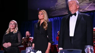 Three formally dressed individuals stand on a stage behind a banquet table with wine glasses, against a blue curtain backdrop.