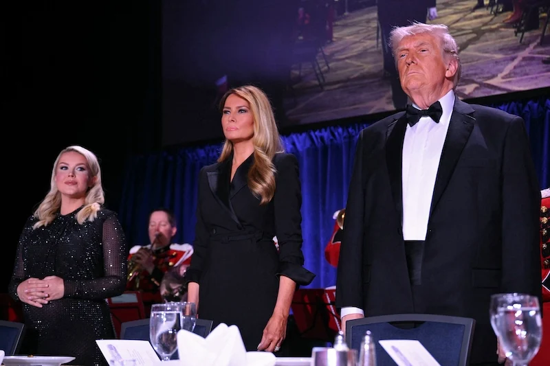 Three formally dressed individuals stand on a stage behind a banquet table with wine glasses, against a blue curtain backdrop.