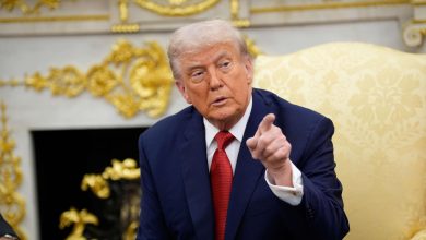 Man in a dark suit and red tie sits in a gold ornate room, gesturing with his finger as he talks.