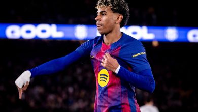 Soccer player in blue and red kit celebrates with outstretched arm as a blurred stadium crowd watches.