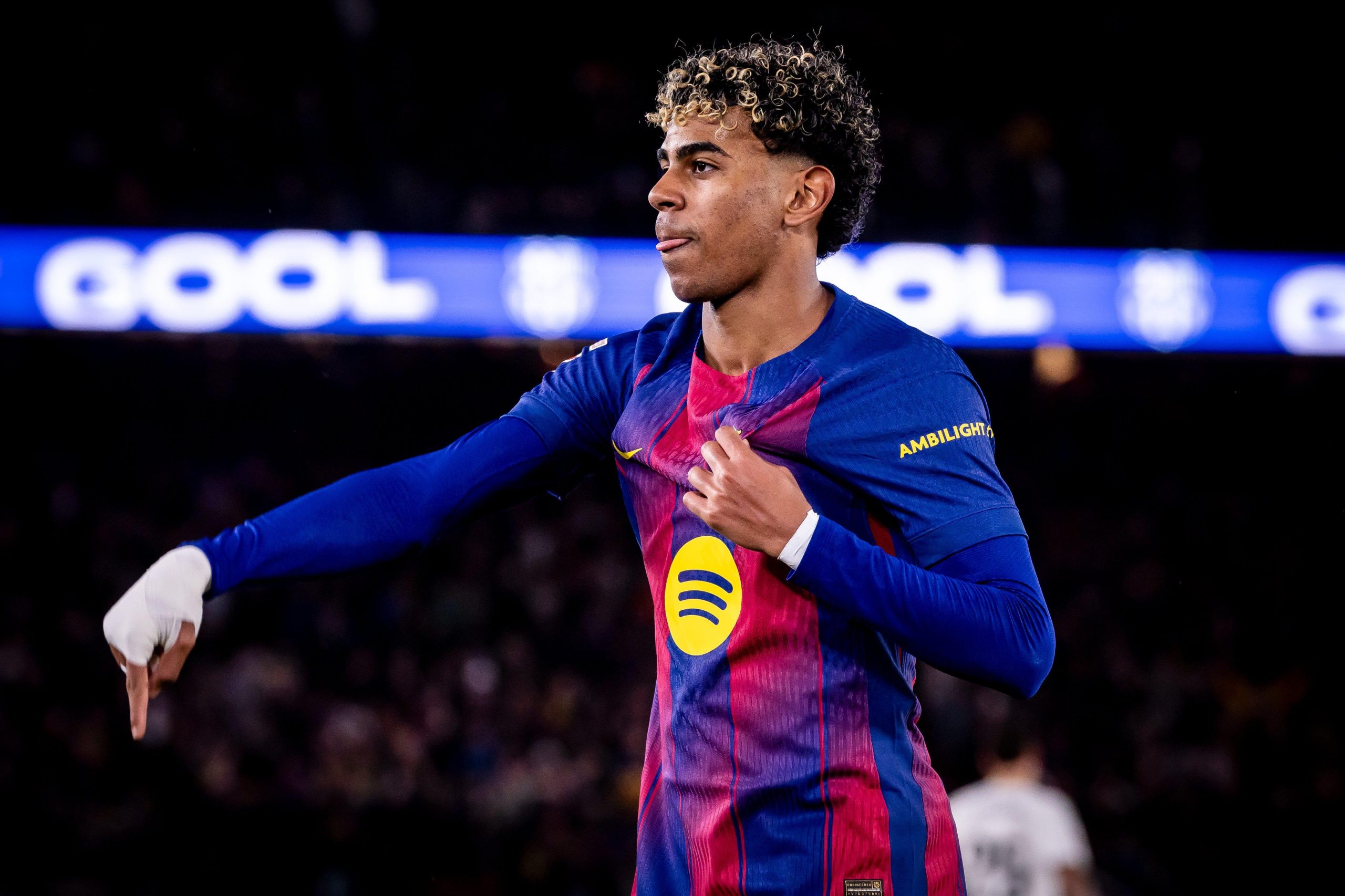 Soccer player in blue and red kit celebrates with outstretched arm as a blurred stadium crowd watches.