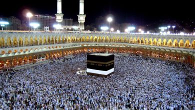 Nighttime view of the Kaaba at Masjid al-Haram, surrounded by thousands of pilgrims and illuminated arched colonnades.