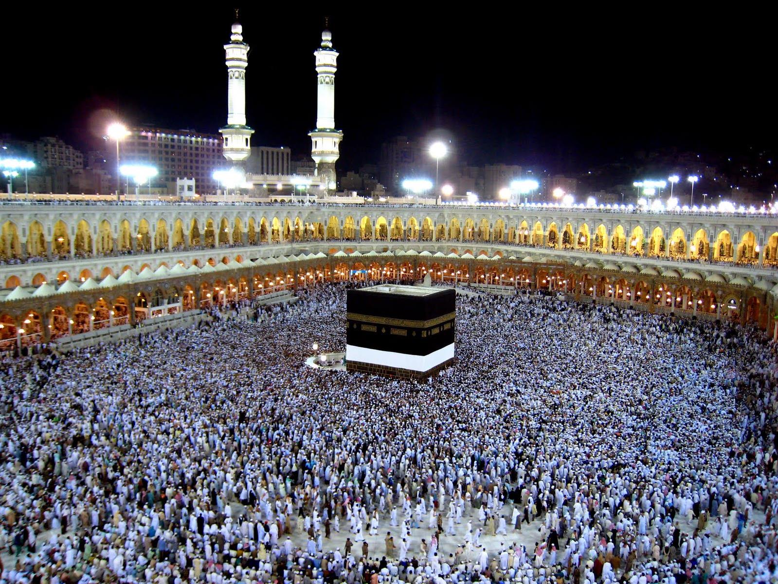 Nighttime view of the Kaaba at Masjid al-Haram, surrounded by thousands of pilgrims and illuminated arched colonnades.