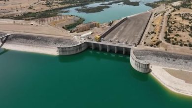 A dam with multiple spillway gates releasing water into a turquoise reservoir, surrounded by arid hills and maintenance equipment on the spillway platform.