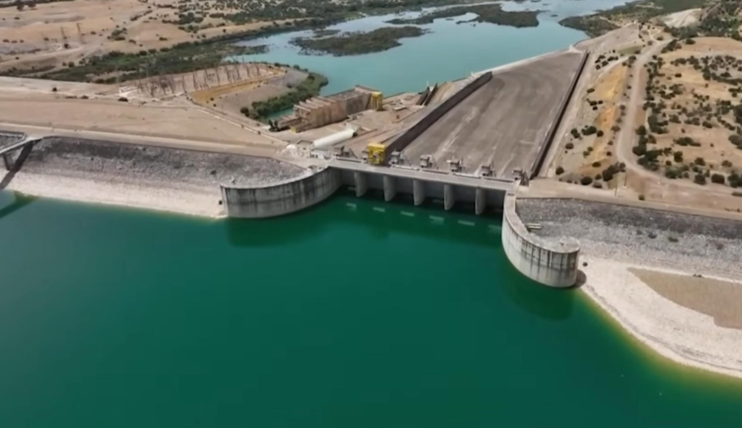 A dam with multiple spillway gates releasing water into a turquoise reservoir, surrounded by arid hills and maintenance equipment on the spillway platform.