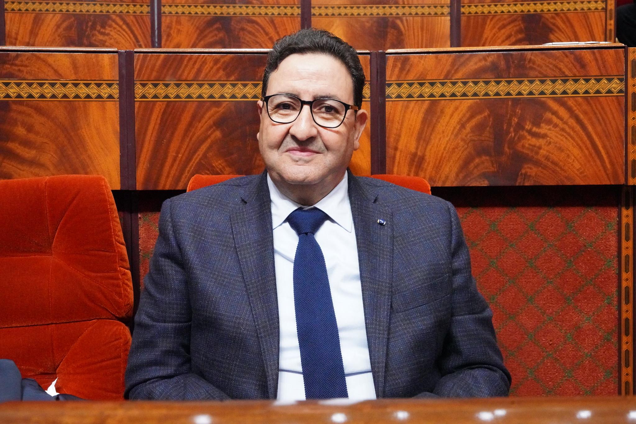 Smiling man in a dark blue checkered blazer, white shirt, and navy tie, seated in a wood-paneled room with red velvet seating and decorative trim behind him.