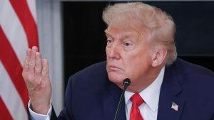 Close-up of a man with blond hair in a dark suit and red tie speaking into a microphone, with an American flag behind him.