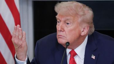 Man in a dark suit and red tie speaks into a microphone, raising his left hand, with an American flag in the background.