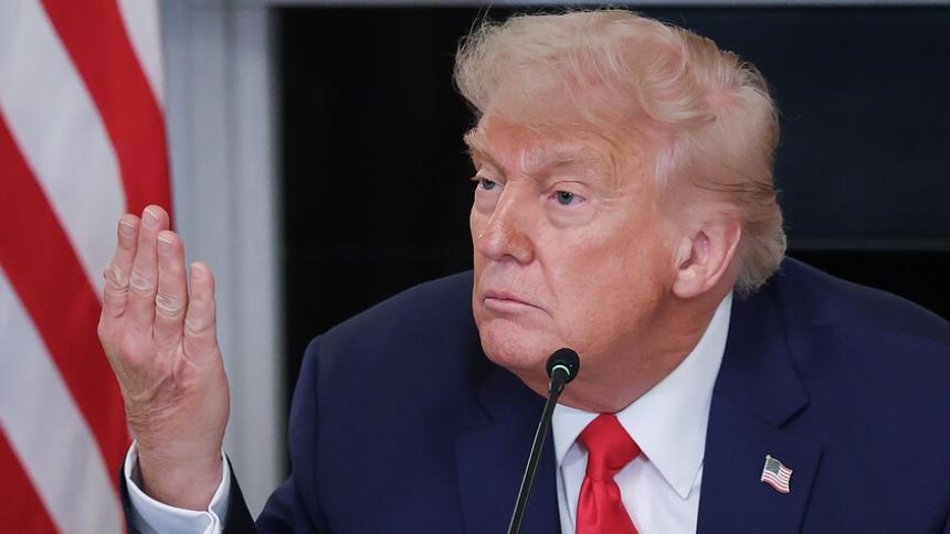 Man in a dark suit and red tie speaks into a microphone, raising his left hand, with an American flag in the background.