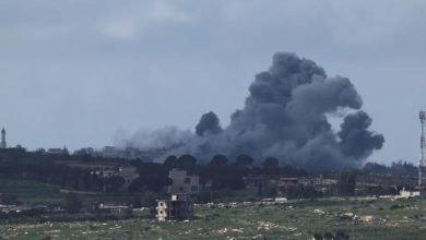 Large black smoke plume rising over a town from an explosion, with scattered buildings and open fields in the foreground.
