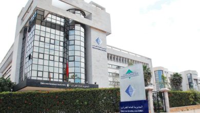 Modern government office building with a glass-front facade, surrounded by hedges and a blue sign in front reading Direction Generale des Impots (GM Tax Office).