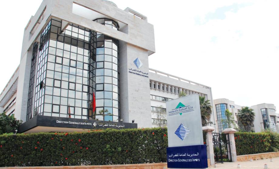 Modern government office building with a glass-front facade, surrounded by hedges and a blue sign in front reading Direction Generale des Impots (GM Tax Office).