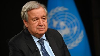 Older man in a dark suit and blue tie sits during an interview, with a United Nations flag blurred in the background.
